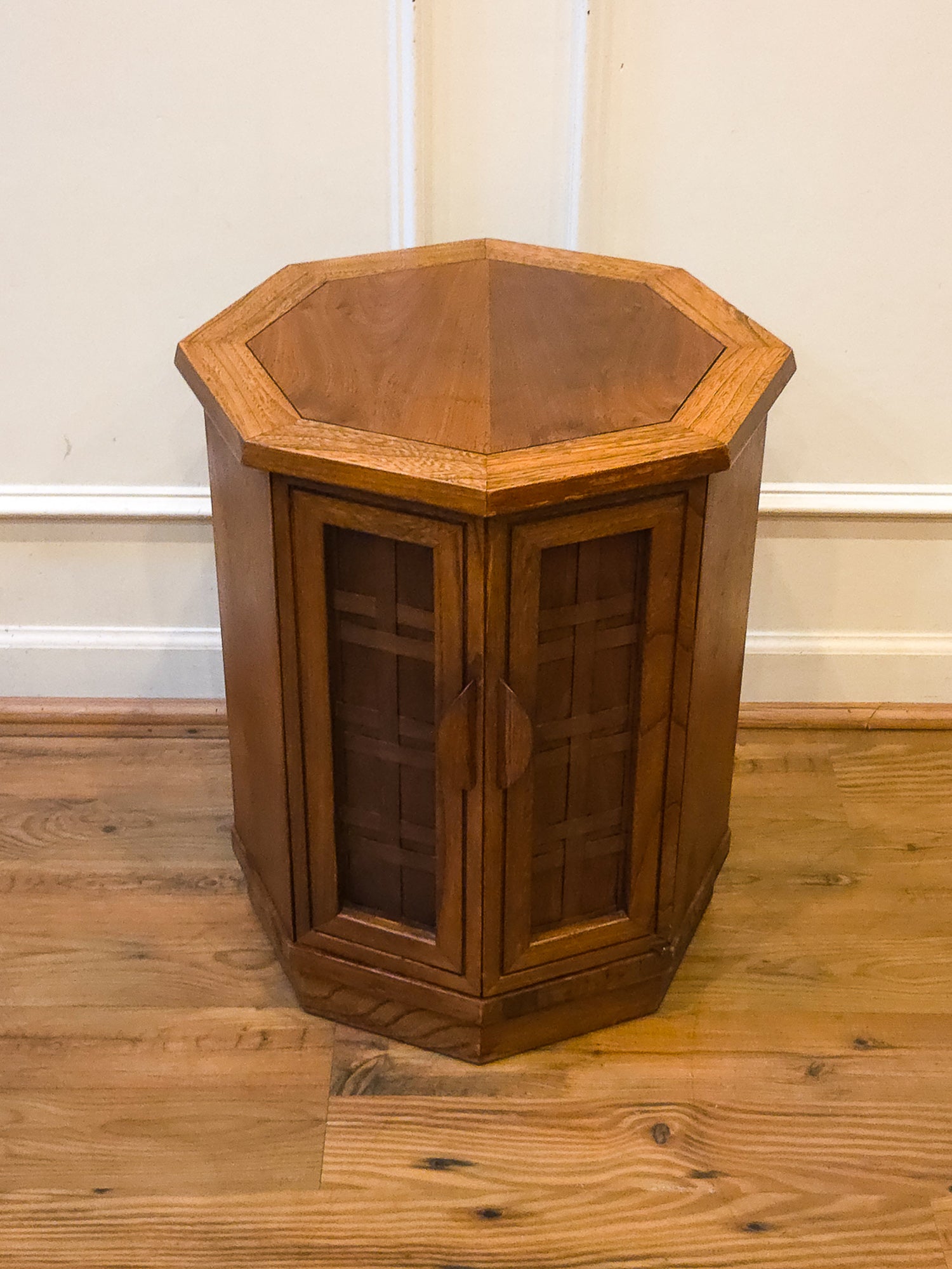 A hexagonal wooden side table or cabinet with glass-paneled doors, shown on a wooden floor against a white wall.