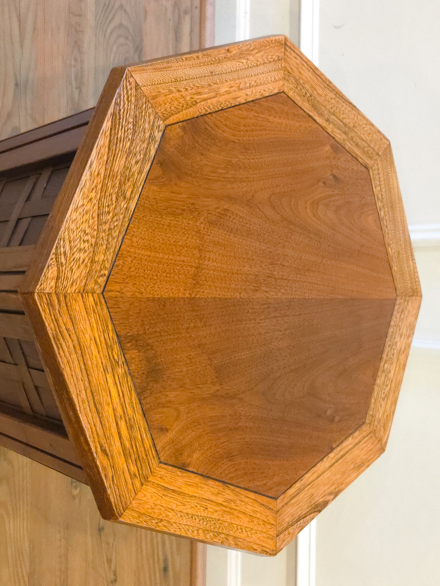 Close-up of an octagonal wooden table top with a warm honey-brown finish and visible wood grain, photographed indoors against a light wall and wood floor.