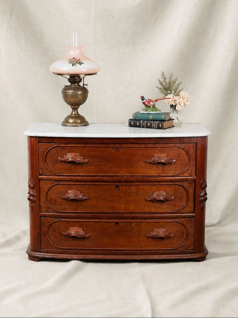 A antique three-drawer wooden chest of drawers with a white marble top, styled with a pink glass oil lamp and a small decorative bird figurine on a draped beige fabric background.