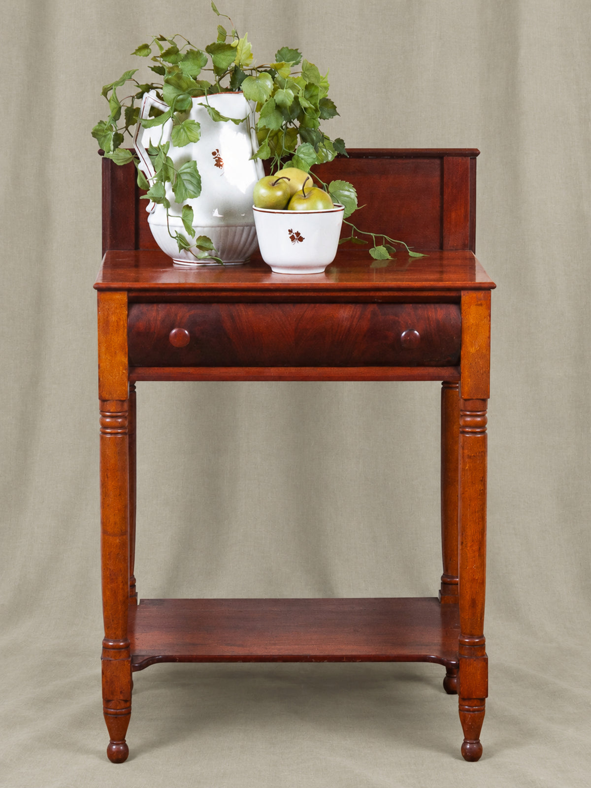 Antique wooden Mahogany wash stand with a single drawer and lower shelf, shown against a neutral beige fabric backdrop. The table is styled with a white ceramic pitcher, green ivy, and a bowl of pears.