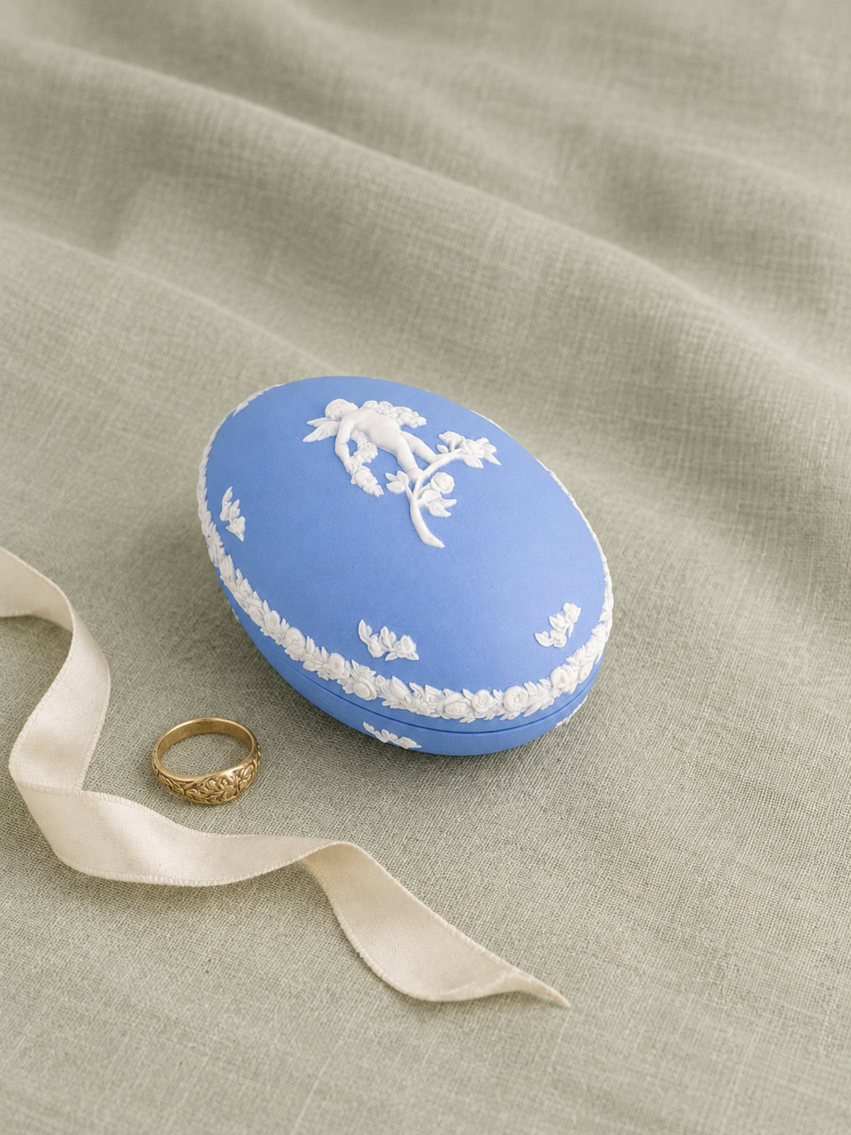 A blue and white Wedgwood-style jasperware ring box with a ribbon loop, placed on a textured sage green fabric background next to a gold ring.