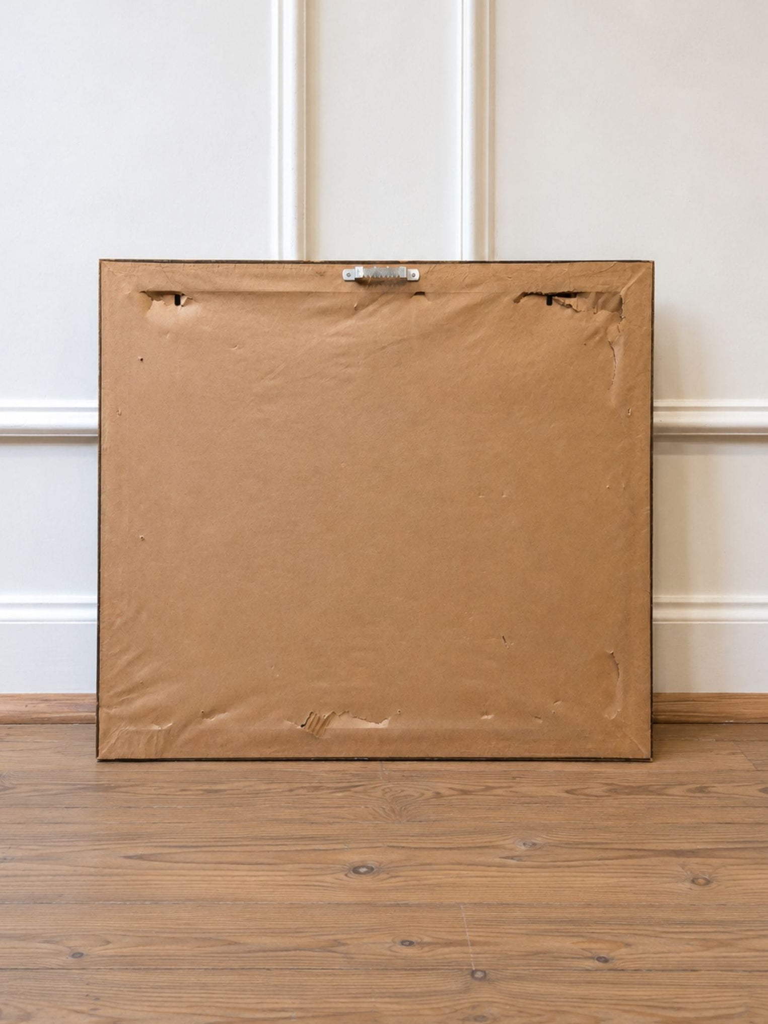 The back of a large square canvas or framed artwork, wrapped in brown paper and secured with a metal clasp, leaning against a white paneled wall on a wooden floor.