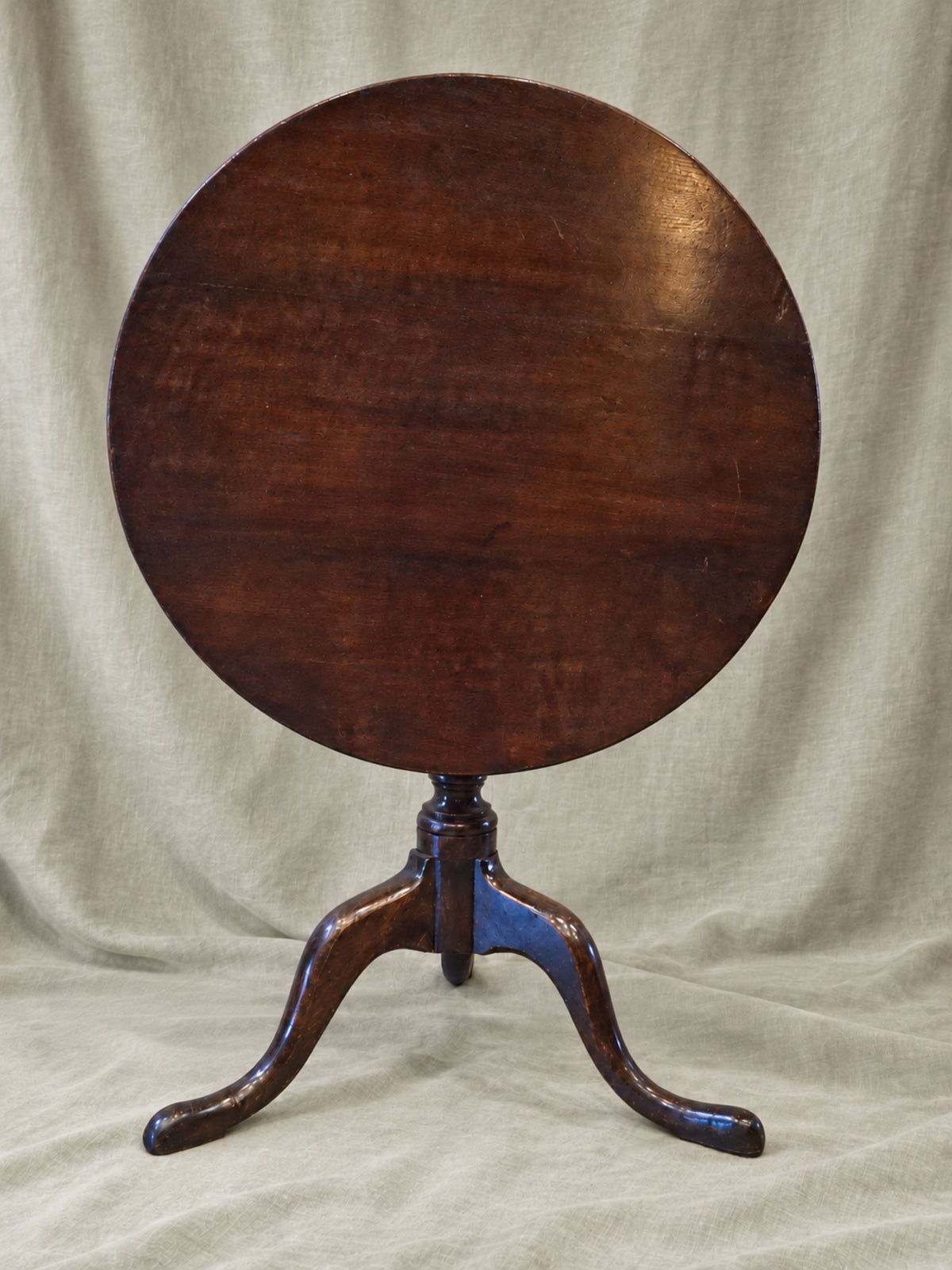 A round wooden tilt-top table with a three-legged pedestal stand, photographed against a draped beige fabric backdrop.