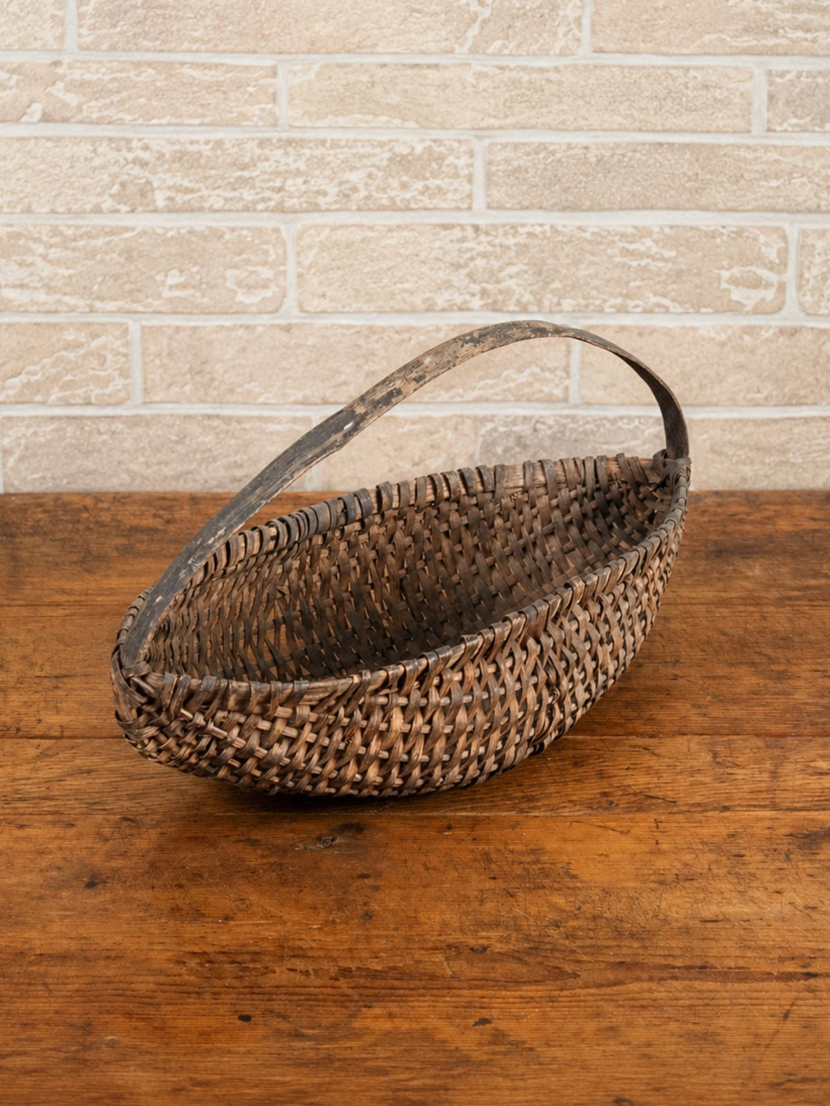 A rustic, hand-woven oval basket with a single loop handle, resting on a wooden table against a light brick wall background.