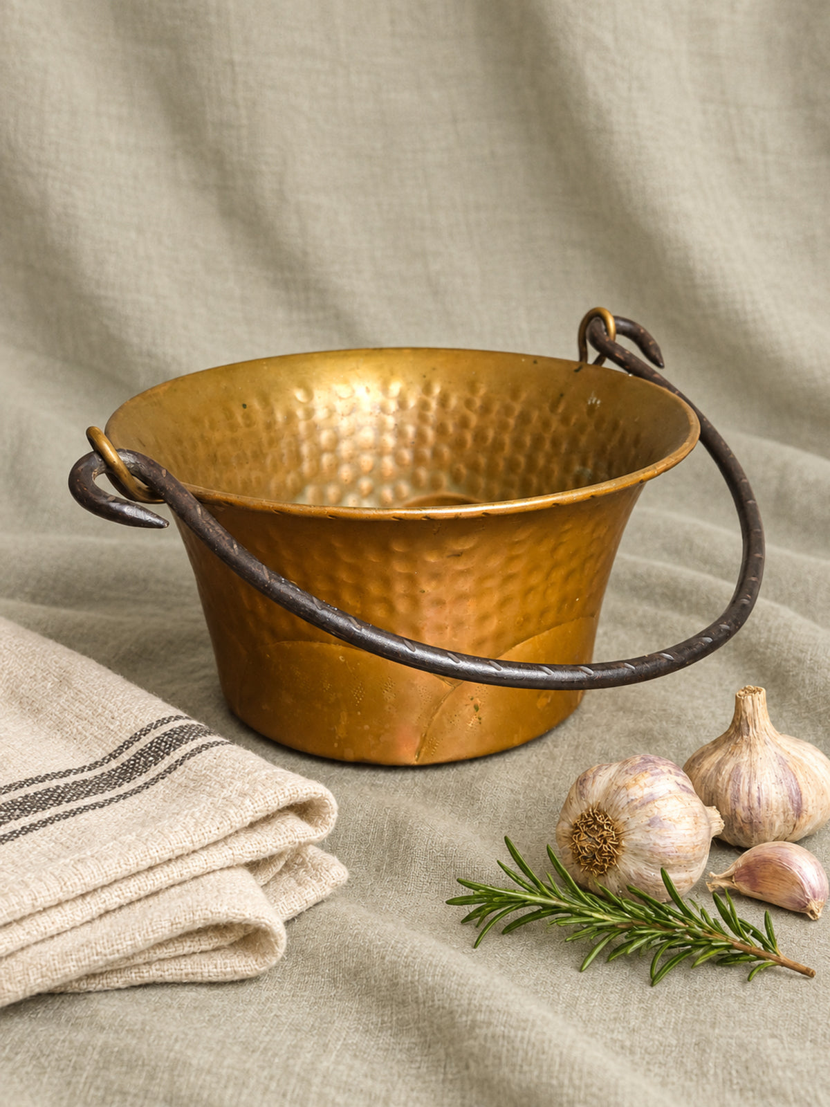 A French hammered copper iron cauldron with a dark metal handle, styled on a beige linen cloth with garlic, a rosemary sprig, and a folded striped towel.