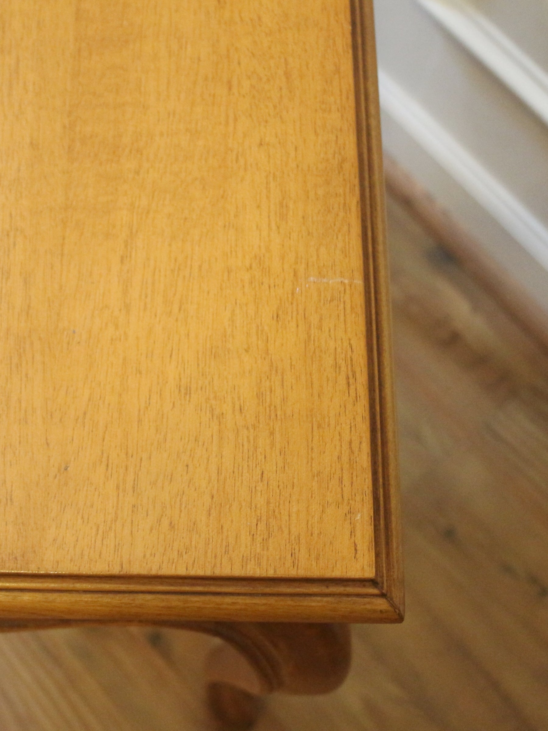 Close-up of a light brown wooden table corner showing the wood grain finish and inner frame edge, with a blurred floor and wall in the background.