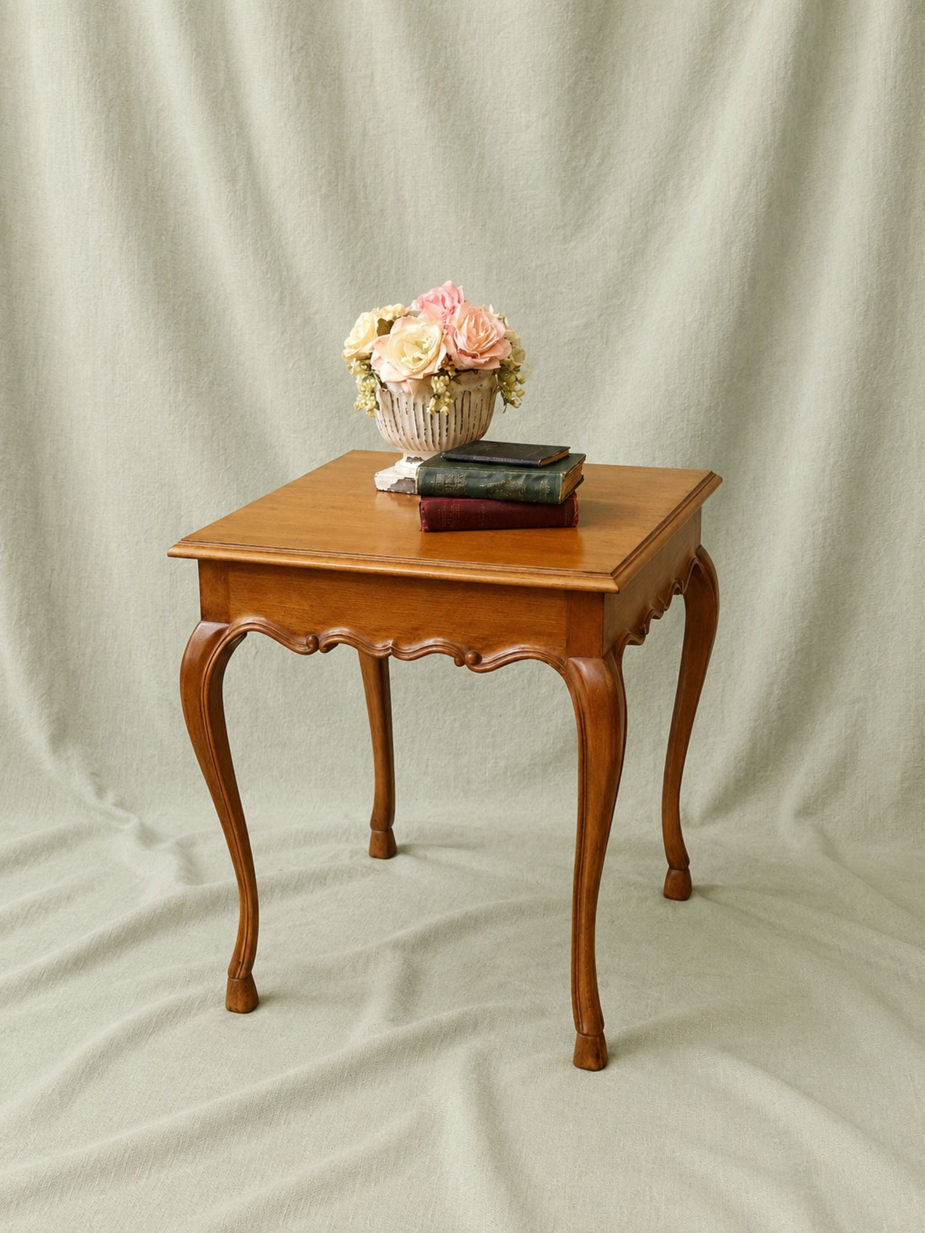 A small square wooden end table with curved cabriole-style legs and a square top, styled with a vase of flowers and stacked books, set against a draped cream fabric backdrop.
