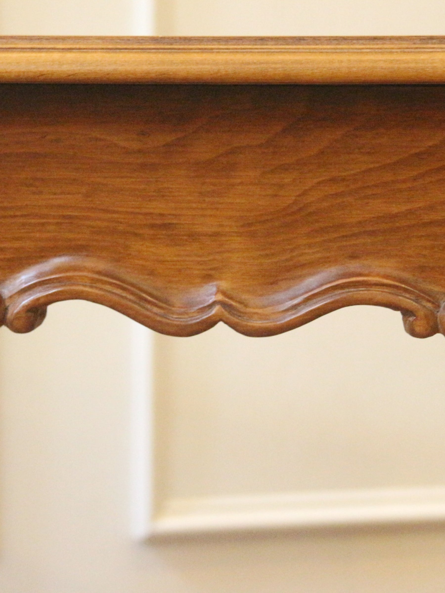 Close-up of a polished medium-brown wooden furniture piece with a carved scalloped apron and visible wood grain, set against a light beige wall.