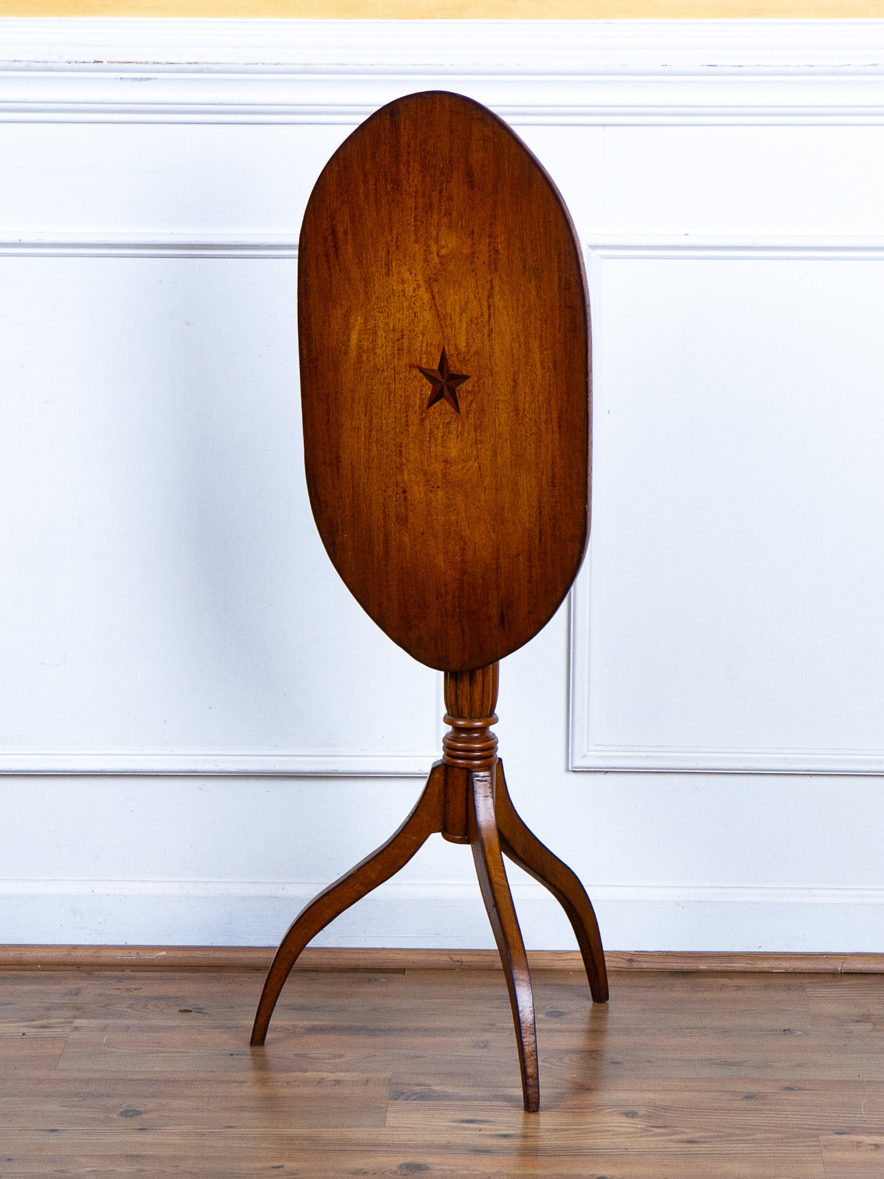 An antique wooden oval tilt-top table on a three-legged tripod stand, featuring a star inlay on the tabletop, set against a white paneled wall and wood floor.