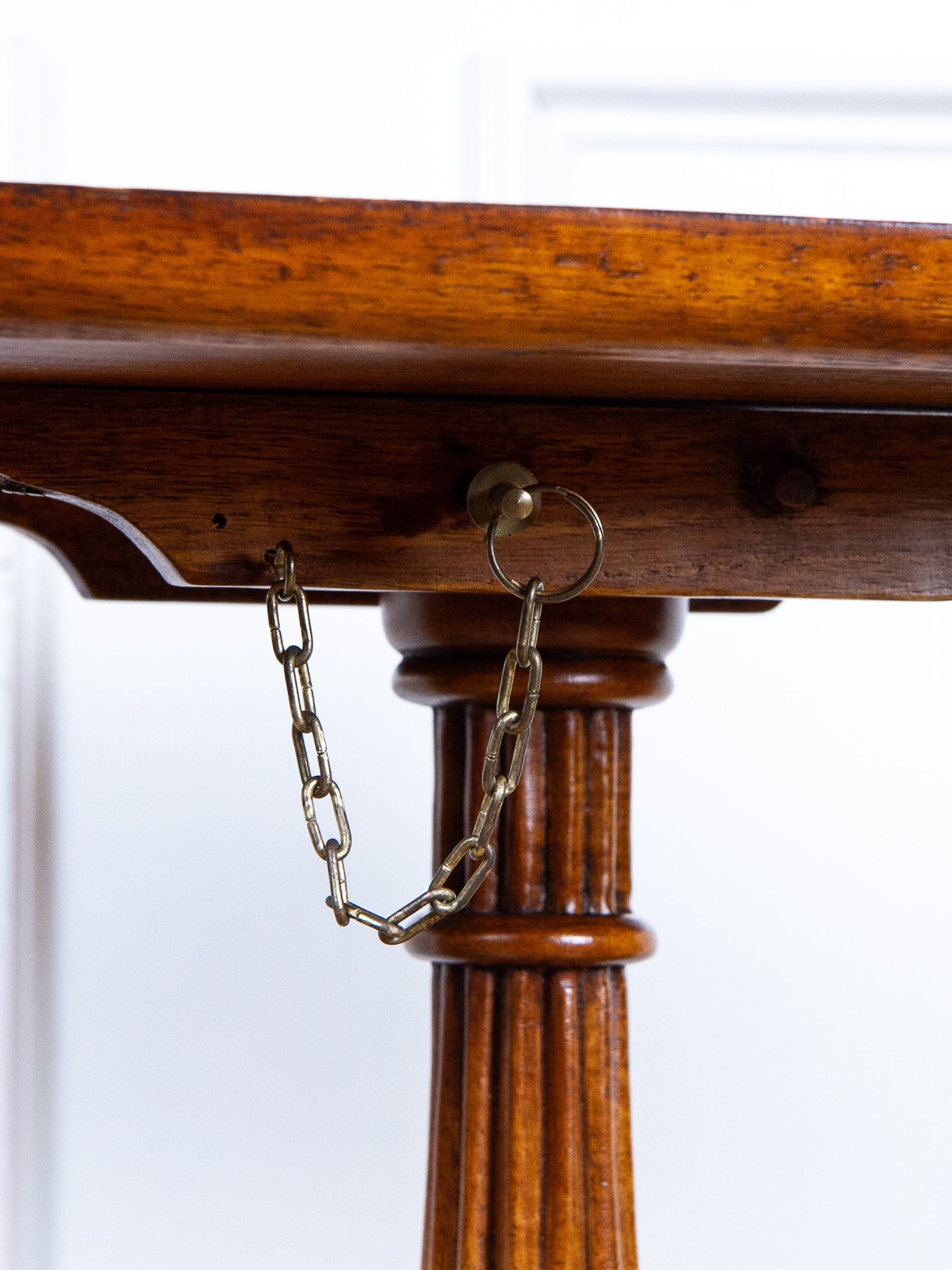Close-up of a wooden tilt-top table with a metal chain attached to a bracket on the tabletop keeping it in place, set against a bright white background.
