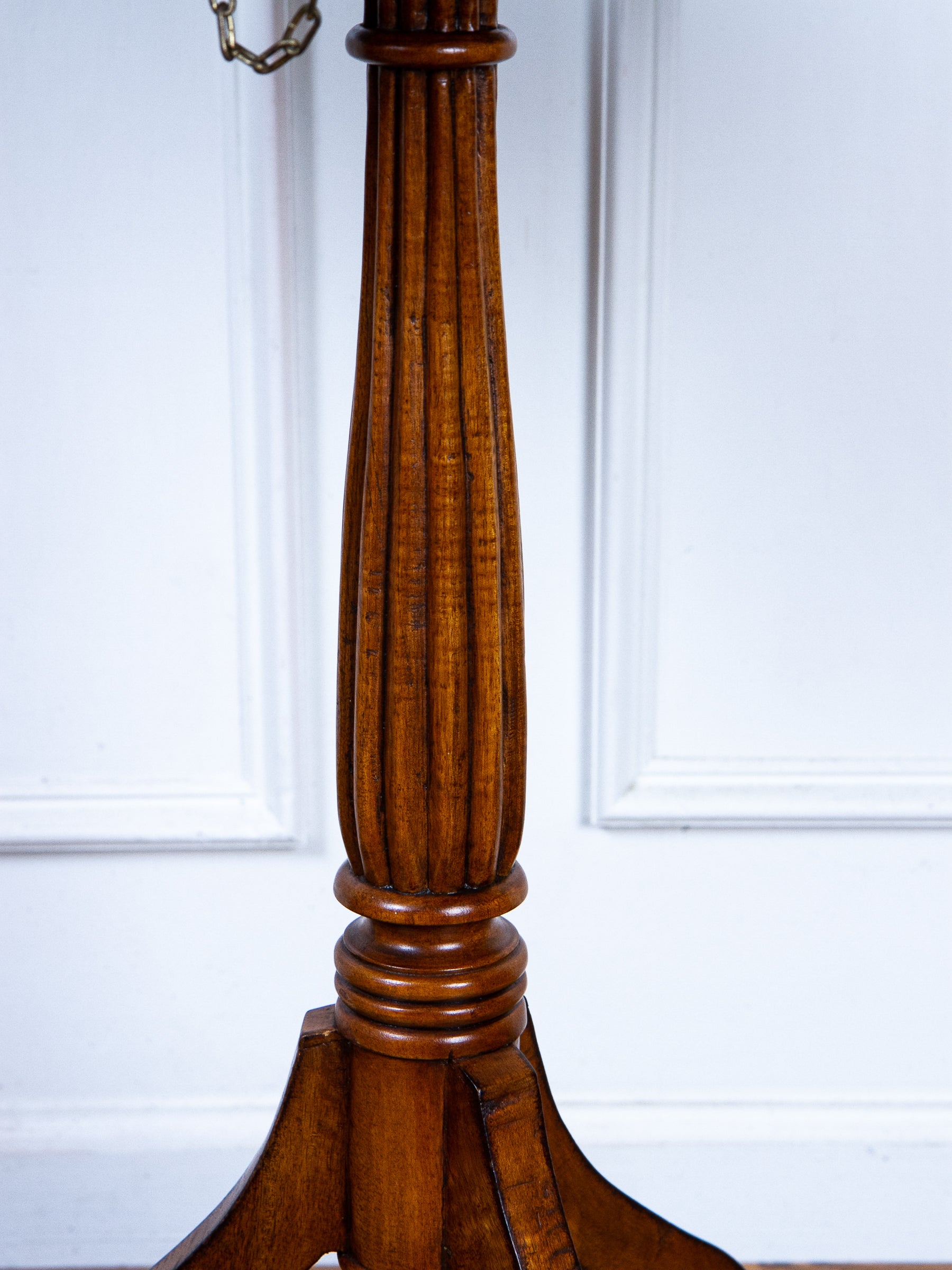 Close-up of a polished dark brown wooden pedestal base with fluted detailing and a chain hanging from the top, set against a white paneled wall.