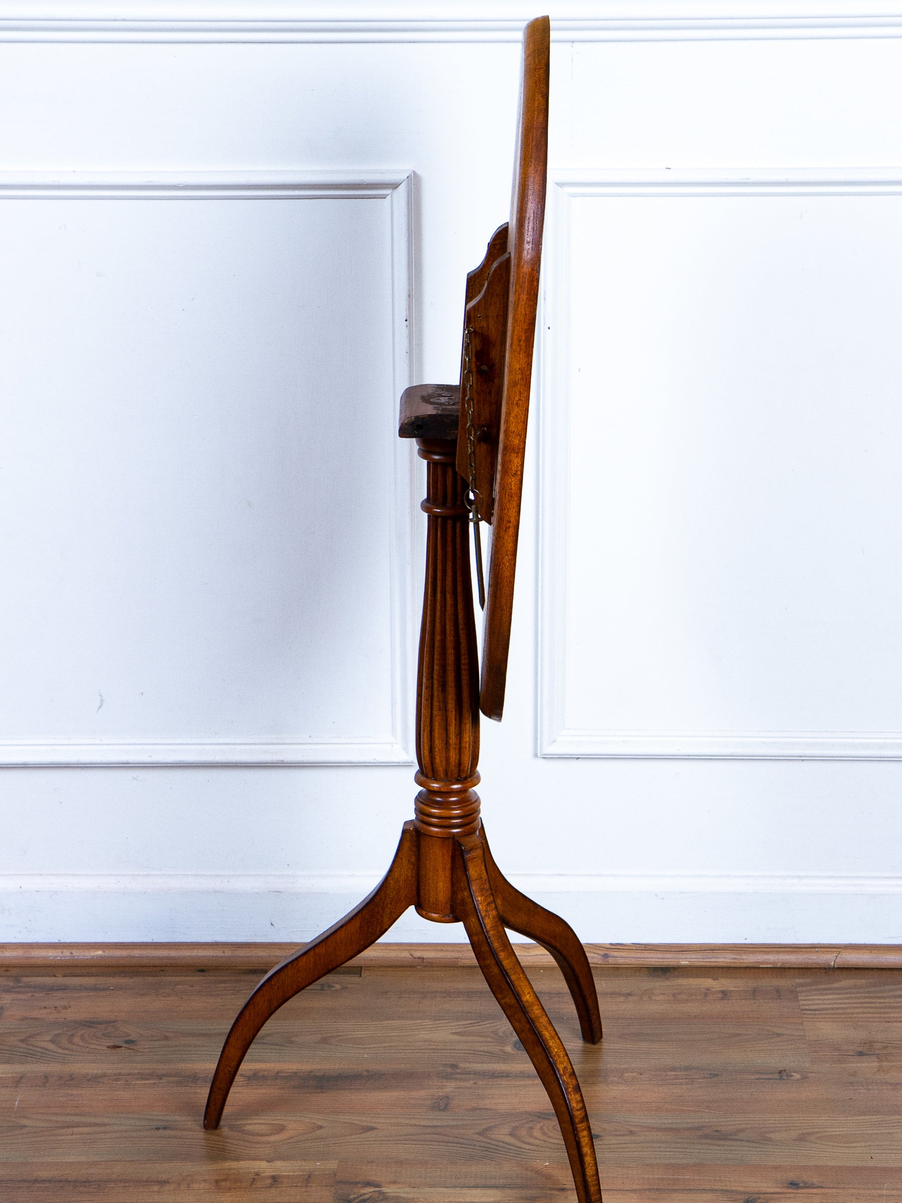 A vintage wooden tilt-top table on a three-legged tripod stand, shown from a side profile view against a white paneled wall and wooden floor.