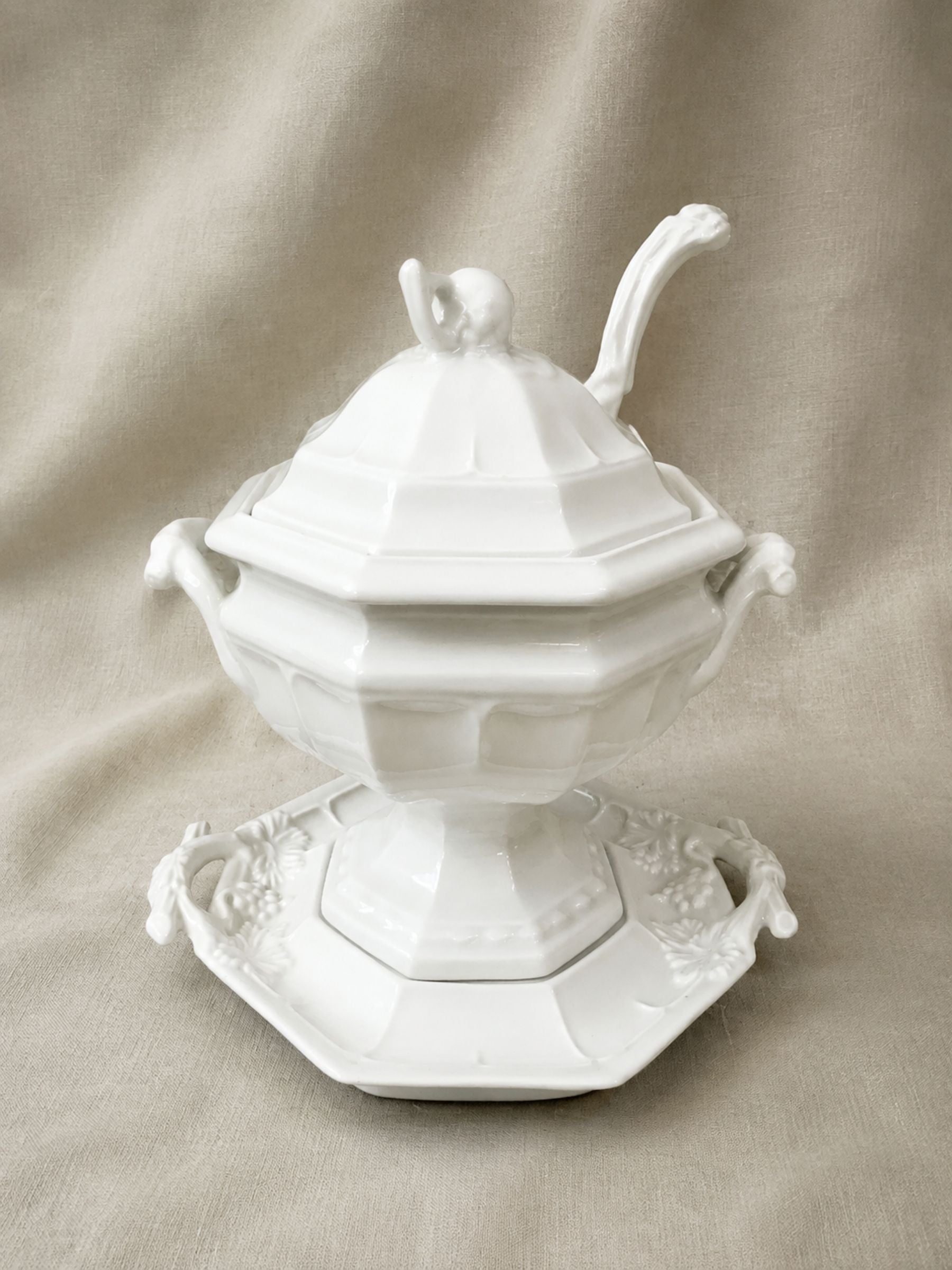 A white porcelain soup tureen  with lid, ladle and decorative side handles and a matching saucer, photographed against a draped beige fabric background.