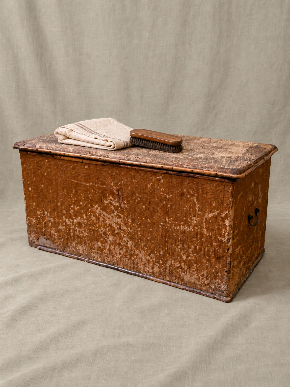 A rustic, weathered brown wooden storage chest with a hinged lid, styled with a folded cream towel and a small wooden brush on top, set against a neutral draped fabric backdrop.